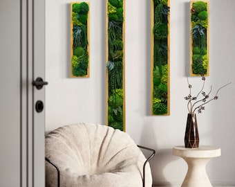 May include: Three vertical framed wall decor pieces with preserved green moss and plants. The frames are made of natural wood and have a light brown finish. The wall decor is displayed on a white wall with a white chair and a small white side table with a vase of dried branches.
