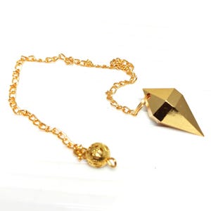May include: A gold-colored metal pendulum with a faceted, geometric, diamond-like point attached to a delicate gold-tone chain. The chain features a small, round, textured decorative bead near the end, creating a simple, metallic design.