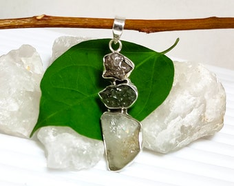 May include: A silver pendant featuring three irregularly shaped stones in shades of gray, green, and white. The pendant is displayed on a green leaf and white background, showcasing the natural textures and colors of the gemstones.