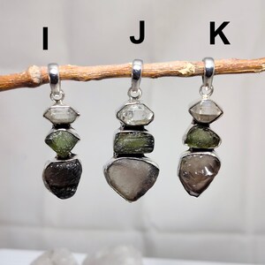 May include: Three silver pendants labelled I, J, and K, each featuring a vertical stack of three raw, irregular gemstones in silver settings. The stones vary in colour, including clear, olive green, and dark grey or brown, suspended from a branch.