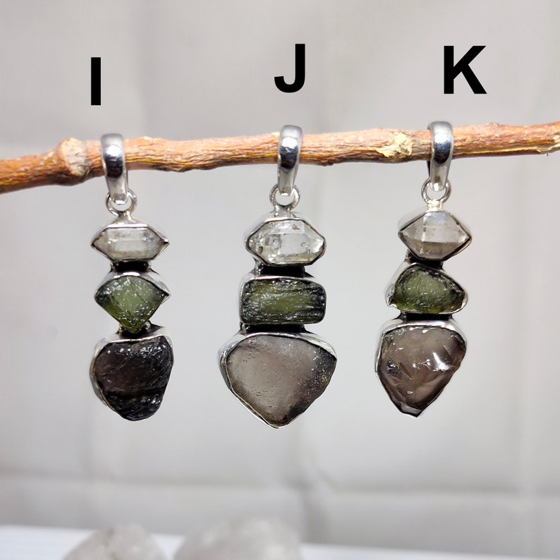 May include: Three silver pendants labelled I, J, and K, each featuring a vertical stack of three raw, irregular gemstones in silver settings. The stones vary in colour, including clear, olive green, and dark grey or brown, suspended from a branch.