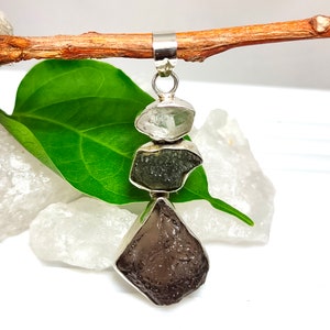 May include: A vertical pendant featuring three stacked, raw, irregularly shaped stones in shades of clear, dark green, and dark brown, each encased in a silver setting with a silver bail at the top for attachment to a chain or cord.
