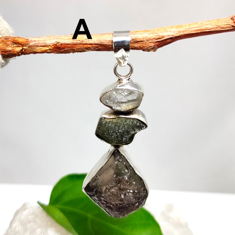 May include: A silver pendant featuring three vertically stacked, raw, irregular gemstones in clear, olive green, and dark brown hues, suspended from a silver bail. The letter A is visible in the upper left corner of the image.