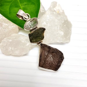 May include: A vertical pendant featuring three raw, irregularly shaped stones set in silver. The top stone is clear, the middle stone is olive green, and the bottom stone is a larger, dark brown piece with a textured, pitted surface finish.