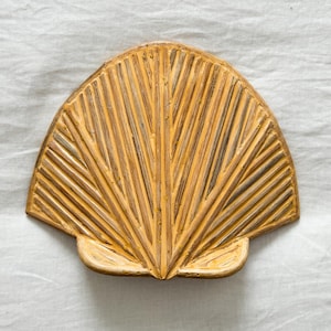 May include: A decorative, fan-shaped object with a textured surface and a warm, golden-yellow hue. The design features parallel lines radiating outwards from a central point, resembling a stylized seashell or palm frond. It is set against a white background.