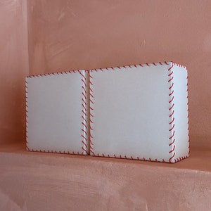 Peut inclure: Deux boîtes rectangulaires blanches avec des coutures rouges autour des bords. Les boîtes sont posées sur une étagère contre un mur rose clair.