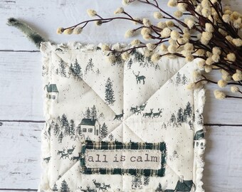 Peut inclure: Un dessous de plat carré et matelassé avec un imprimé de scène hivernale. Le tissu crème présente une forêt, des cerfs et des maisons dans des tons de gris et de vert. L'expression "all is calm" est imprimée sur un patch à carreaux verts.