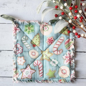 May include: A square, quilted Christmas potholder with a light blue background and various appliqued cookie designs. The cookies include gingerbread men, candy canes, stars, and snowflakes. The potholder has a ruffled edge and a hanging loop.
