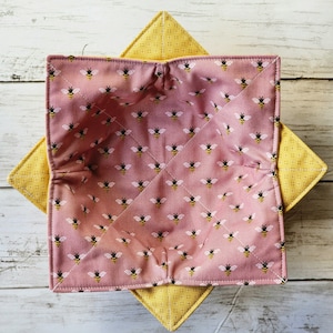 May include: A pink fabric bowl cozy with a repeating pattern of black and yellow bees. The cozy is quilted with white thread and sits atop a yellow square with a small white dot pattern.