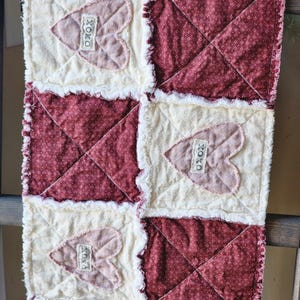May include: A handmade, quilted table runner featuring alternating squares of cream and burgundy fabric. Cream squares have heart appliqués with "XOXO" labels. The burgundy squares have a small white polka dot pattern. The edges are frayed.