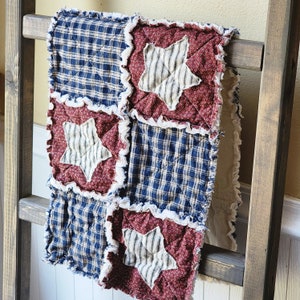 May include: A red, white, and blue patchwork quilt with a frayed edge and stars in the center of each square. The quilt is hanging on a wooden ladder.
