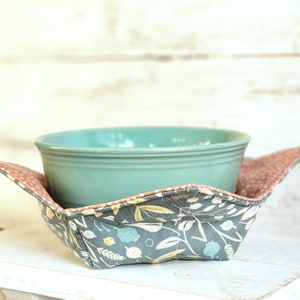 May include: A teal ceramic bowl sits inside a gray and white floral fabric bowl cozy. The cozy has a brown and white polka dot trim.