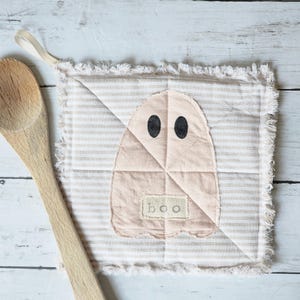 May include: A beige and white striped square pot holder with a stitched ghost design. The ghost has black oval eyes and a small tag that says "boo". The pot holder has a frayed edge and a loop for hanging.