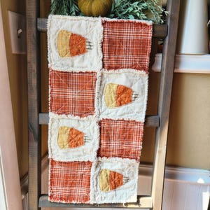 May include: A handmade, patchwork table runner featuring a repeating pattern of candy corn and orange plaid squares. The candy corn is yellow, orange, and white, with a frayed edge. The runner is displayed on a wooden ladder.