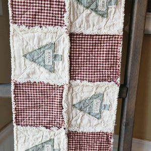 May include: A quilted Christmas tree runner with a patchwork design. The runner features alternating squares of red and white gingham and off-white fabric. Each off-white square has a green tree applique with the word "merry". The edges have a frayed finish.