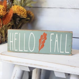 May include: A rectangular wooden sign with the words "HELLO FALL" in white lettering. An orange leaf design is between the words. The sign has a light blue-green background and sits on a white surface.