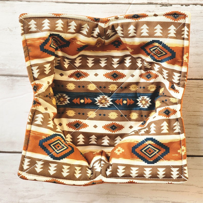 Peut inclure: Un panier en tissu matelass&eacute; brun et orange avec un motif g&eacute;om&eacute;trique. Le panier est doubl&eacute; d'un tissu blanc et a une forme carr&eacute;e.