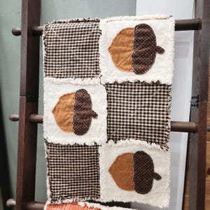 May include: A rag quilt featuring a patchwork design of alternating squares. Some squares have a black and white gingham check pattern, while others are off-white with appliqued brown and tan fabric acorns with frayed edges.