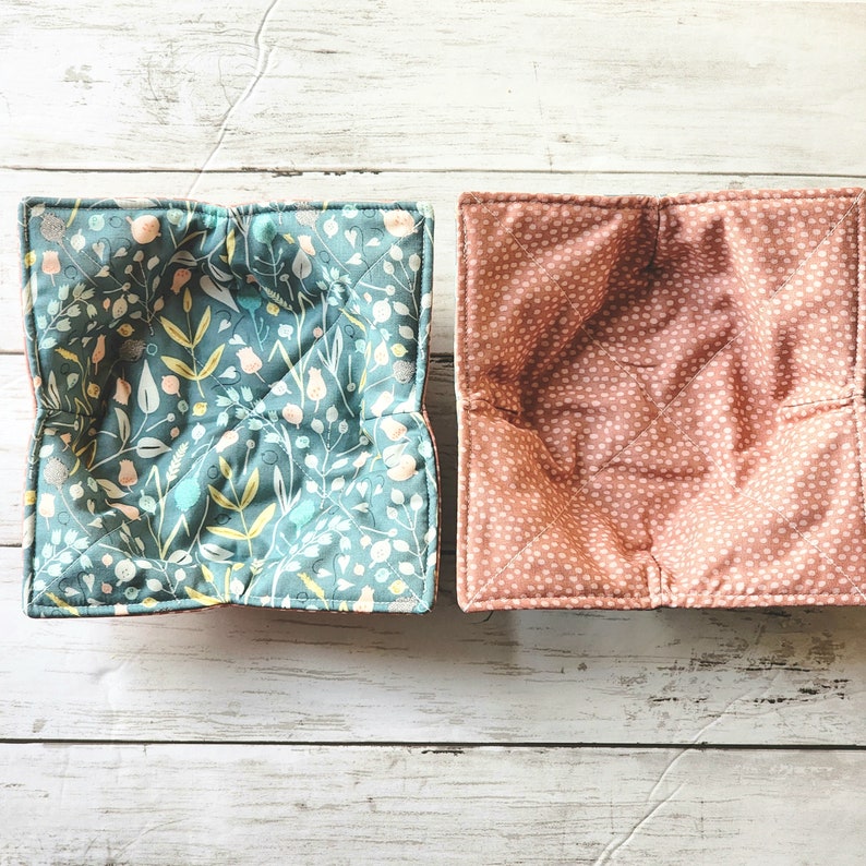 May include: Two fabric bowl cozies, one with a floral pattern in shades of blue and green, and the other with a pink and white polka dot pattern.