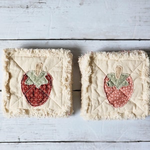 May include: Two square, cream-colored fabric coasters with a stitched strawberry design. The strawberries are red and pink with green leaves and tan stems. The edges of the coasters have a frayed, fringed trim.