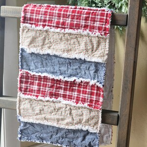 May include: A patchwork quilt with alternating stripes of red plaid, beige floral, and blue fabric. The edges of each fabric strip are frayed, creating a textured, rustic appearance. The quilt is draped over a wooden ladder.