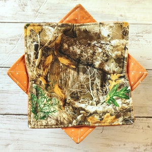 May include: Square fabric hot pad with a camouflage pattern, featuring shades of brown, green, and tan. The hot pad is layered over an orange square with white polka dots, creating a contrasting design.