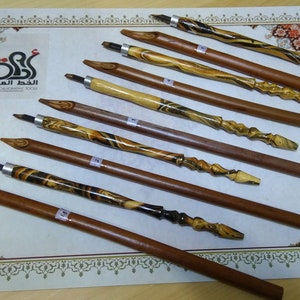 Könnte beinhalten: Ein Satz von zehn Kalligraphie-Stiften aus Holz mit verschiedenen Holzmaserungen. Die Stifte haben Metallspitzen und sind auf einer weißen Oberfläche mit einem dekorativen Rand angeordnet. Der Text "الخط العربي" ist in der oberen linken Ecke des Bildes sichtbar.