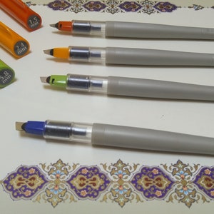 May include: A collection of calligraphy pens with gray barrels and colored tips. The pens have different tip sizes, indicated by the colored caps: orange (1.5 mm, 2.4 mm), green (3.8 mm), and blue (6.0 mm). The pens are arranged on a decorative surface.