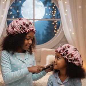 May include: Two young people wearing matching pink bonnets and light blue pajamas with white star patterns. One is brushing the other's hair. A plush bunny and open books are on the floor. The scene is set in a cozy bedroom with a large window and fairy lights.