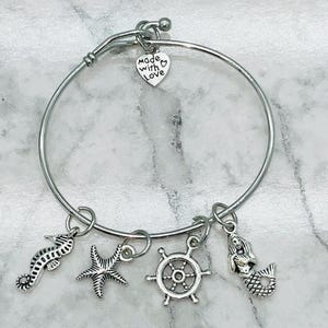 May include: Silver-toned bangle bracelet with nautical charms. The bracelet features a seahorse, starfish, ship's wheel, and mermaid charm. A heart-shaped charm with the words "made with love" is also included. The bracelet is displayed on a marble surface.