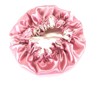 May include: A pink satin bonnet with a cream-colored interior. The bonnet is gathered at the edges with an elastic band, creating a ruffled effect. The shiny fabric reflects light, highlighting the soft pink hue.