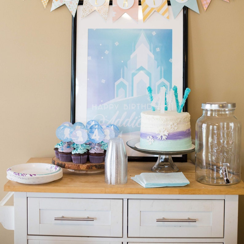 May include: A white birthday cake with blue and purple frosting and snowflake decorations. The cake is on a white cake stand and has a sign that says "Happy Birthday Addie". There are cupcakes with blue and purple frosting and snowflake decorations on a wooden tray. There is a clear glass dispenser with a silver lid and a silver tap. There are also blue and white paper napkins and a silver plastic cup on the table.