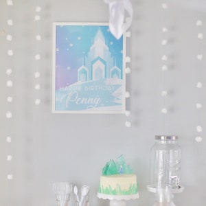 May include: A white table with a blue and white cake decorated with a small figurine and green icing. The cake is on a white cake stand. There is a clear glass dispenser with a silver lid and a blue and white plate on the table. The background is a light blue wall with white pom-poms hanging from the ceiling. A framed poster with a blue and white castle and the text "Happy Birthday Penny" is on the wall.