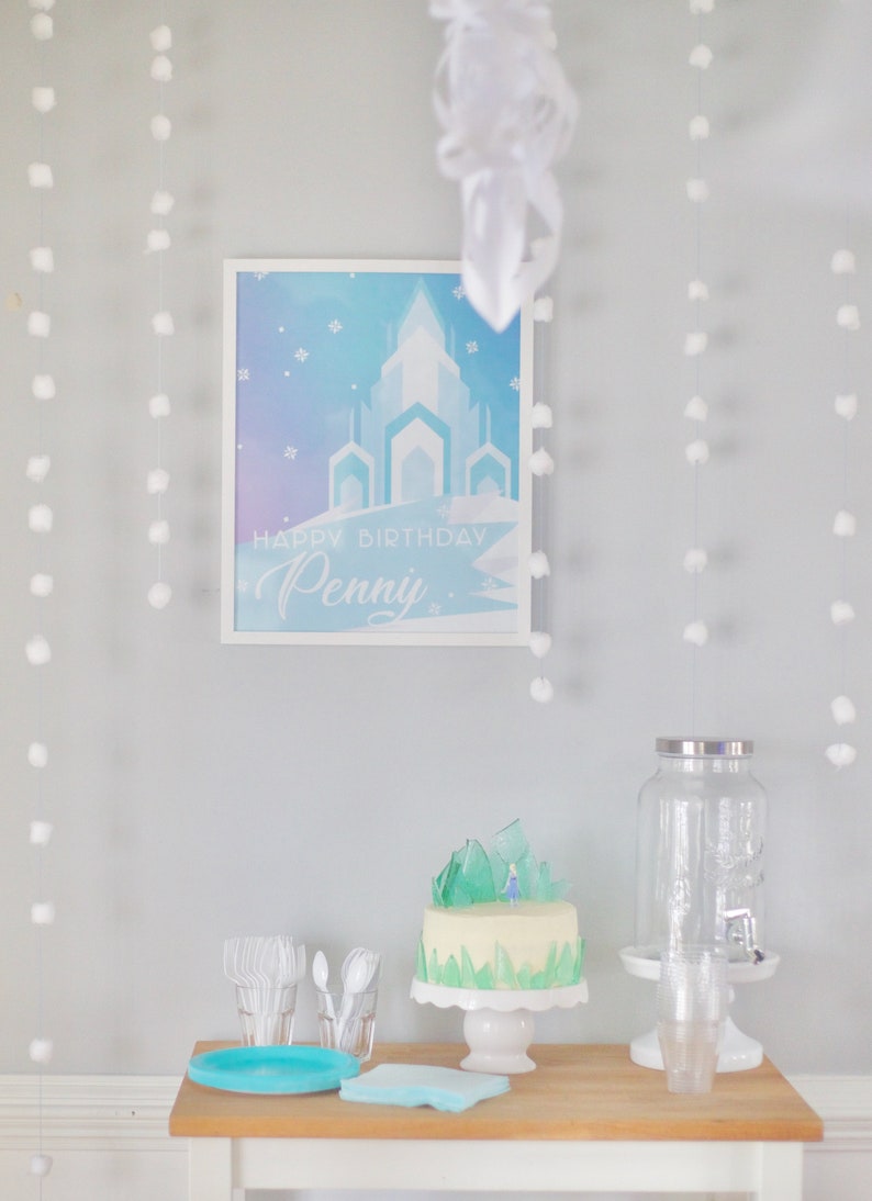 May include: A white table with a blue and white cake decorated with a small figurine and green icing. The cake is on a white cake stand. There is a clear glass dispenser with a silver lid and a blue and white plate on the table. The background is a light blue wall with white pom-poms hanging from the ceiling. A framed poster with a blue and white castle and the text "Happy Birthday Penny" is on the wall.