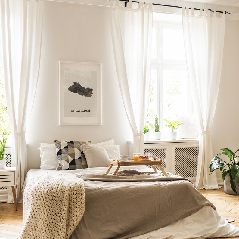 May include: A bedroom with white walls and white curtains. A bed with a brown blanket and white pillows is in the centre of the room. A framed print of the country of El Salvador is hanging on the wall behind the bed. A tray with a glass of orange juice and a plate of fruit is on a wooden tray table next to the bed.