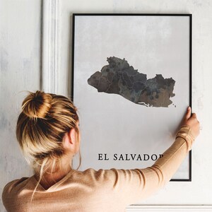 May include: A black and white print of a map of El Salvador, with the country name printed below the map. The print is framed in a black frame and is being hung on a white wall by a person.