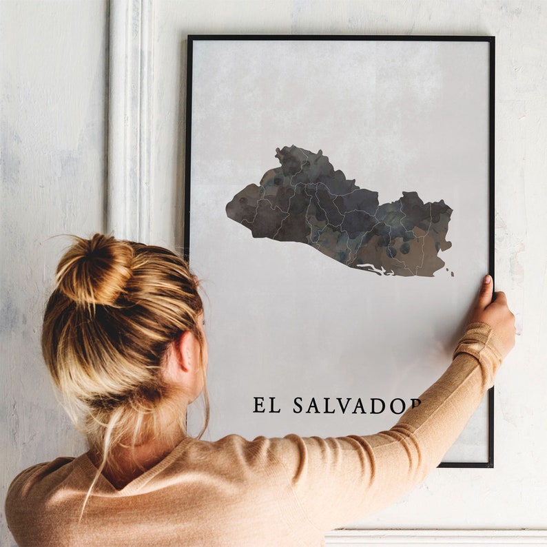May include: A black and white print of a map of El Salvador, with the country name printed below the map. The print is framed in a black frame and is being hung on a white wall by a person.