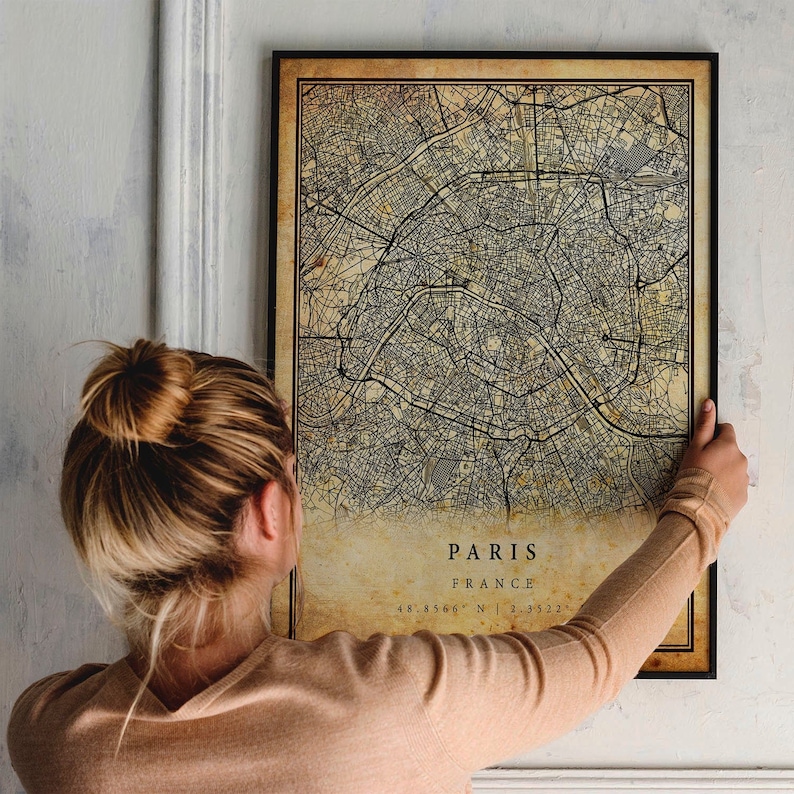 May include: A vintage-style map of Paris, France, printed on aged paper with a black frame. The map shows the city's streets and landmarks. The text "Paris, France 48.8566&deg; N | 2.3522&deg; E" is printed at the bottom of the map.