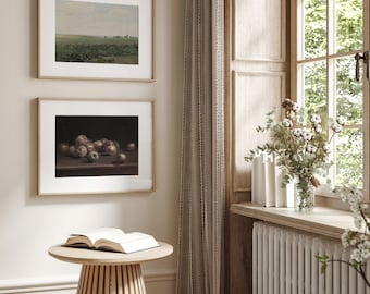 May include: Two framed landscape and still-life paintings hang on a wall. The top painting depicts a field under a cloudy sky. The bottom painting shows apples on a table. A small, round wooden table holds an open book.