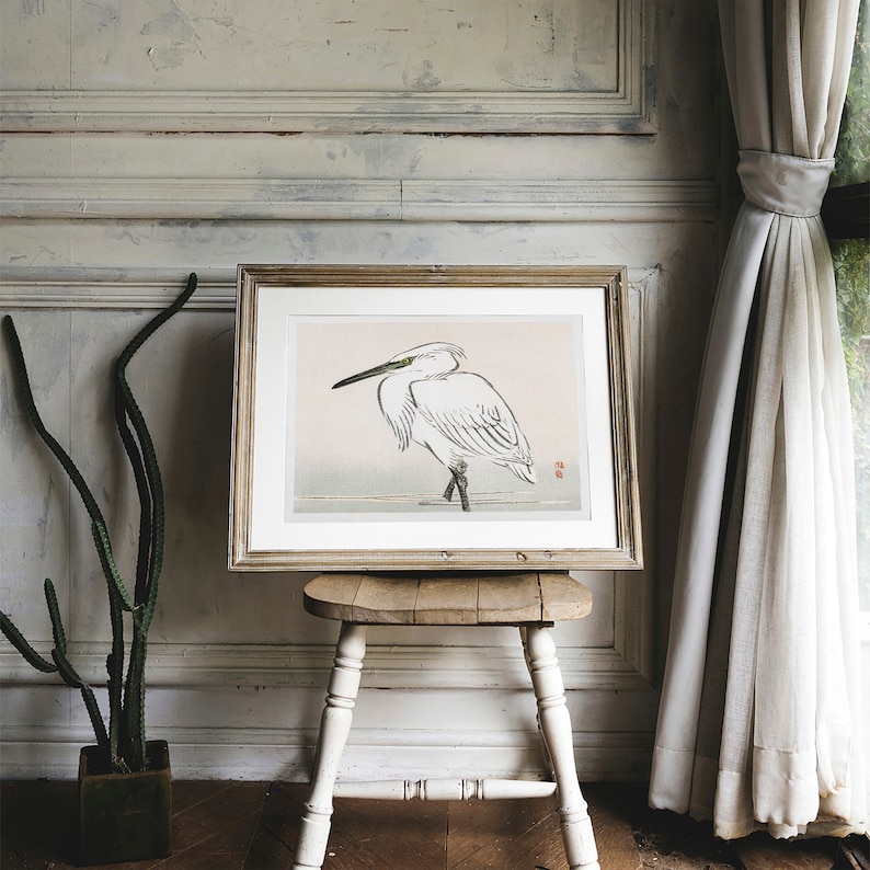 May include: A framed print of a white egret standing on one leg with its head turned to the side. The print is on a light pink background with a subtle gray wash. The frame is a simple, light wood.