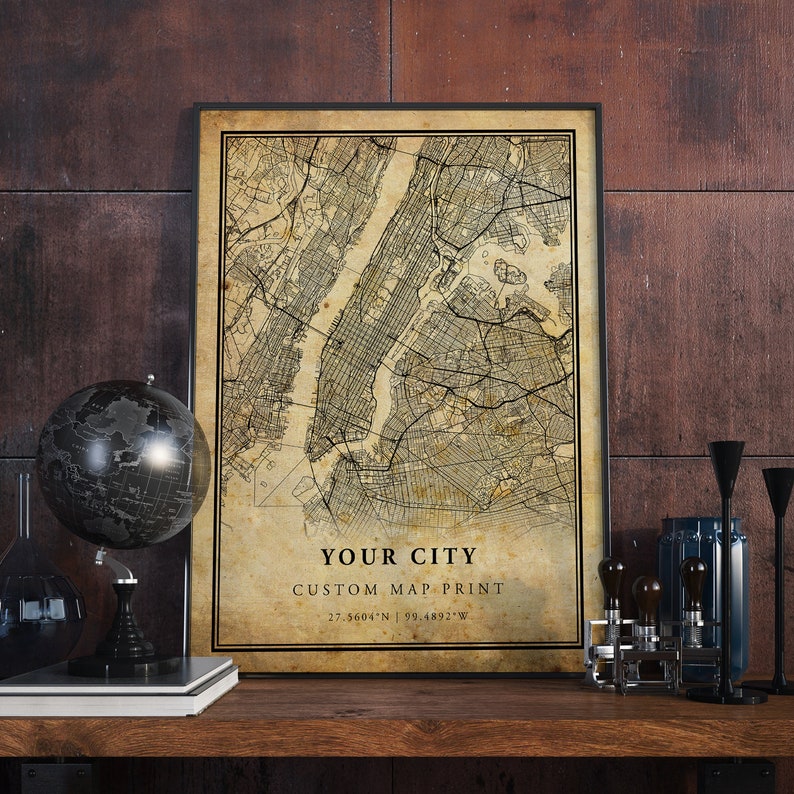 May include: A vintage-style map print of a city, framed in black. The map is in shades of brown and beige, with black lines for streets and waterways. The text "YOUR CITY CUSTOM MAP PRINT 27.5604&deg;N 99.4892&deg;W" is printed at the bottom of the map.