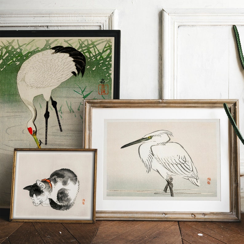 May include: Three framed prints of Japanese woodblock prints. The top print features a white crane with black markings standing in a green and brown landscape. The middle print features a white egret with black markings standing in a gray and white landscape. The bottom print features a black and white cat curled up sleeping.