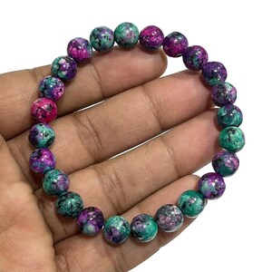 May include: A beaded bracelet featuring round beads in shades of green, purple, and pink. The bracelet is held in a hand, showcasing the colors and textures of the gemstones. The beads appear to be approximately 0.4 inches in diameter.