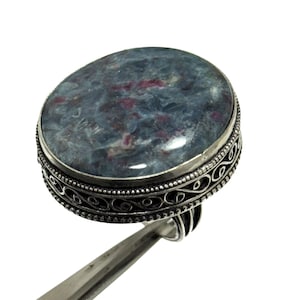 May include: A ring featuring a large, oval-shaped, polished blue and red mottled gemstone set in a silver band. The silver setting displays intricate filigree scrollwork patterns along the side, complemented by a beaded border detail.
