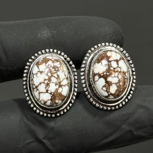 May include: A pair of oval-shaped earrings featuring white and brown patterned stones set in a silver-toned metal frame. The stones have a distinct mottled appearance, and the metal border is decorated with a small, raised beaded texture.