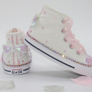 May include: White high-top Converse sneakers with pink ribbon laces and a sparkly pink bow on the side. The shoes are covered in iridescent rhinestones.