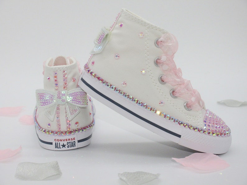 May include: White high-top Converse sneakers with pink ribbon laces and a sparkly pink bow on the side. The shoes are covered in iridescent rhinestones.