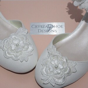 May include: A pair of white satin bridal shoes with delicate lace flower embellishments. The shoes have a low heel and a simple design.