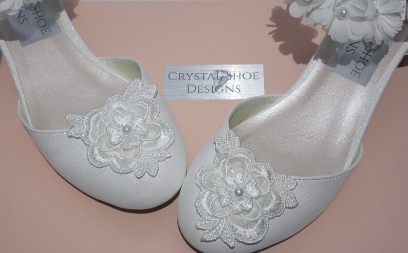 May include: A pair of white satin bridal shoes with delicate lace flower embellishments. The shoes have a low heel and a simple design.