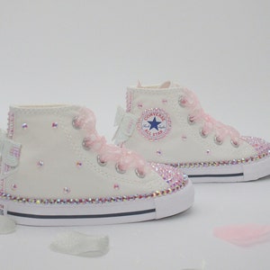 May include: White high-top Converse sneakers with pink ribbon bows and rhinestones. The sneakers have a blue star logo and the text "Converse All Star" on the side.